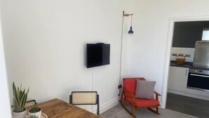 Ett kompakt vardagsrum med ett matbord i trä, en röd vadderad stol, en väggmonterad TV och en köksdel med en fläktkåpa i rostfritt stål.