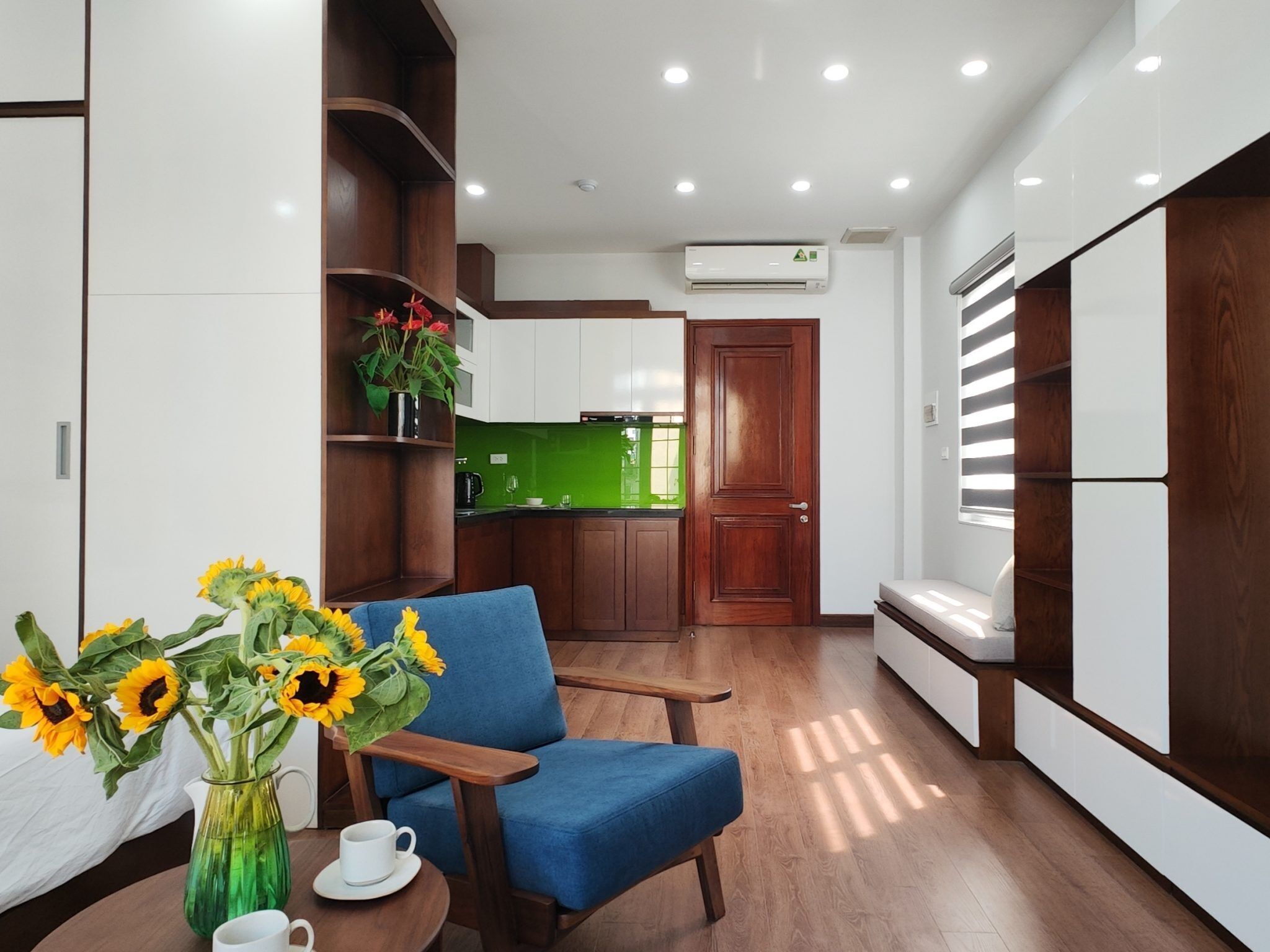 Une cuisine moderne avec des meubles blancs, un pare-feu vert et un plancher en bois. On y trouve une chaise bleue et un vase avec des tournesols.