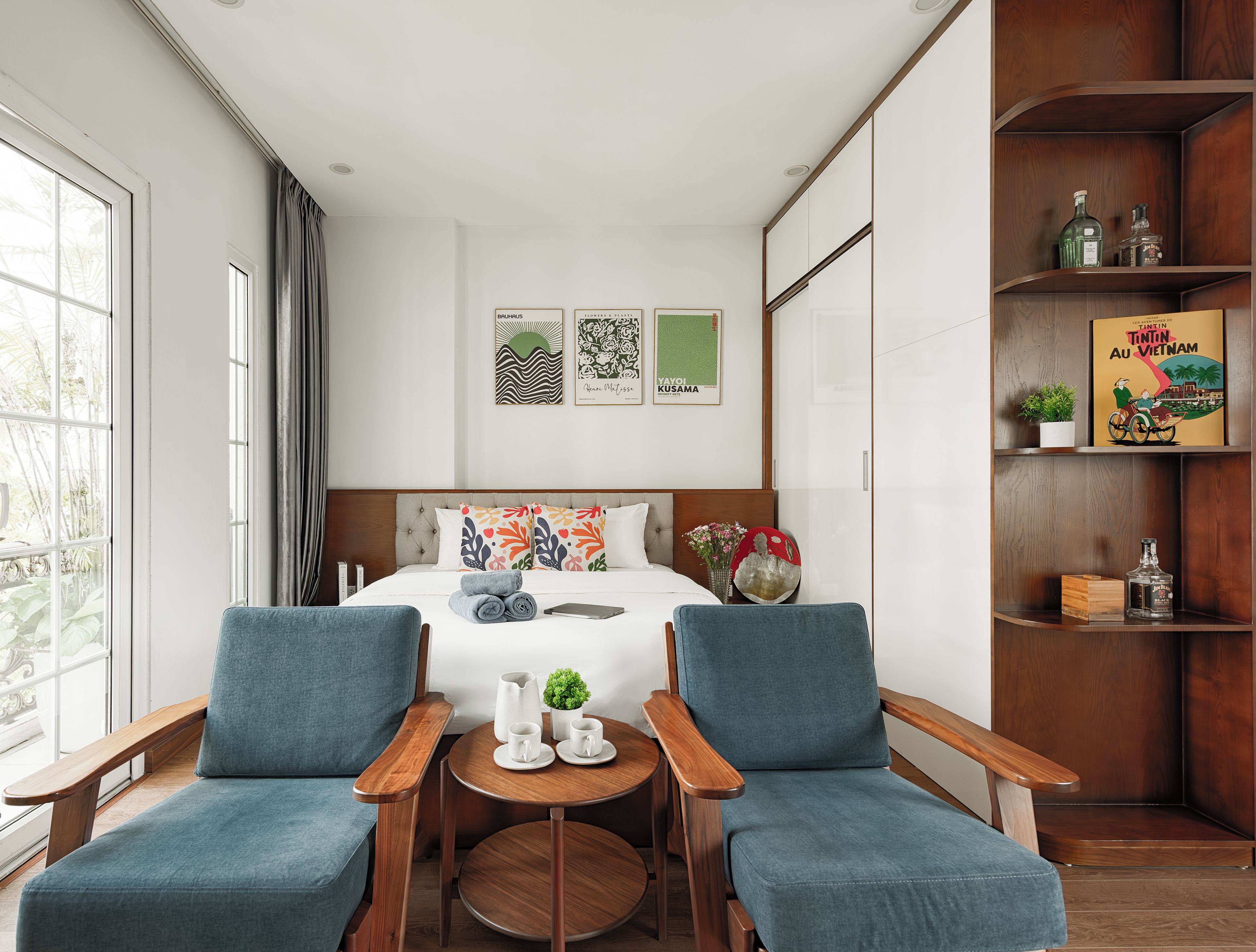 Una habitación de hotel con una cama, dos sillones, una mesita con una planta y una estantería llena de libros y objetos decorativos.