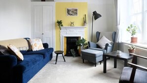A living room with a blue sofa, a grey armchair, a small table, a fireplace, and a window with curtains. - Fantastic 2BD Victorian Apt - 4 adults and 1 child (East Sussex)