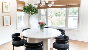 A dining area with a round table, six chairs, a vase with flowers, a hanging light fixture, and large windows with a view of greenery. - New! Chic Cottage Home W/ Backyard Pool - 12 min to the River Walk! (Alamo Heights)