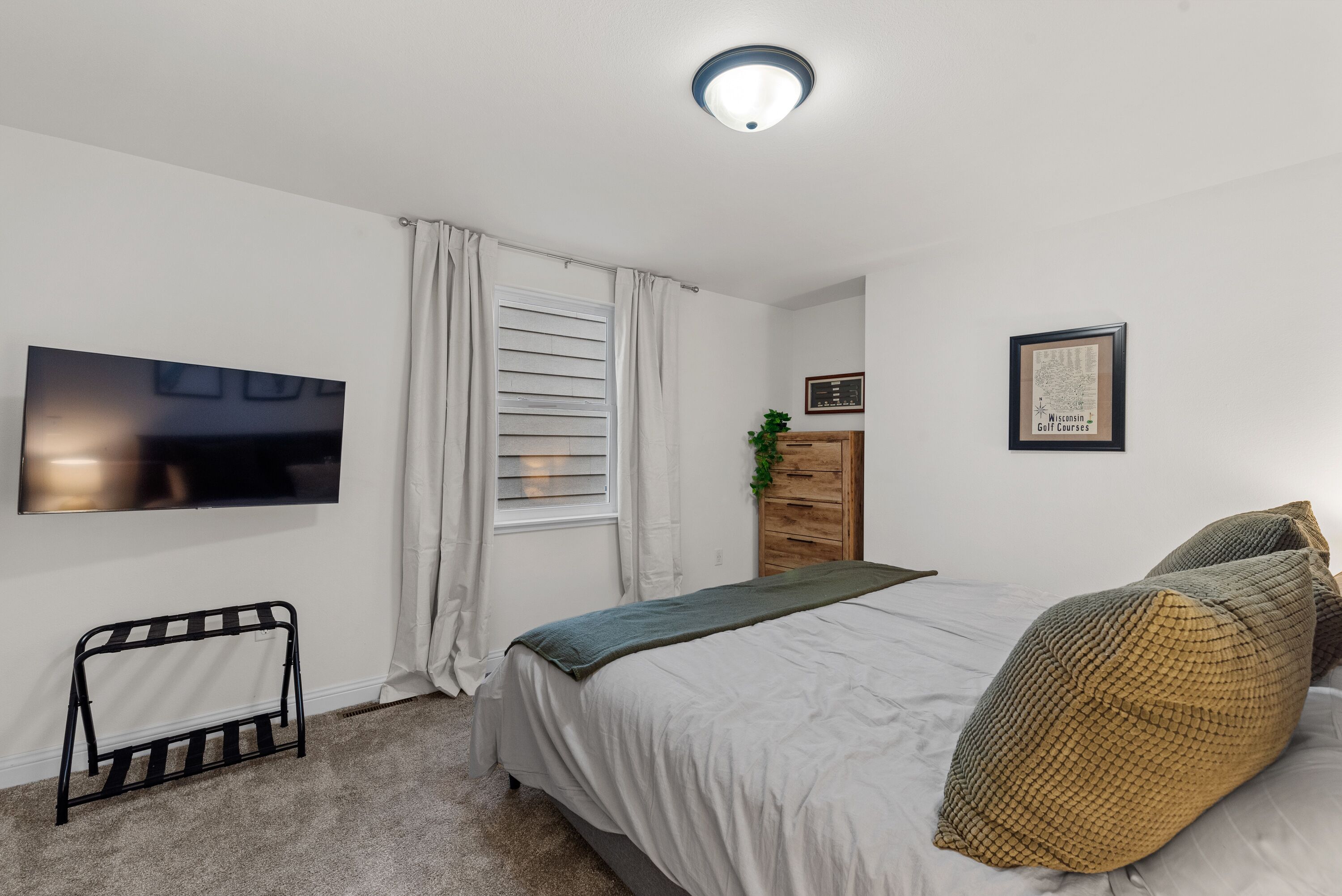 Een slaapkamer met een bed, een aan de muur bevestigde televisie, een raam met gordijnen, een dressoir en een ingelijste foto aan de muur.