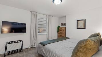 Een slaapkamer met een bed, een aan de muur bevestigde televisie, een raam met gordijnen, een dressoir en een ingelijste foto aan de muur.