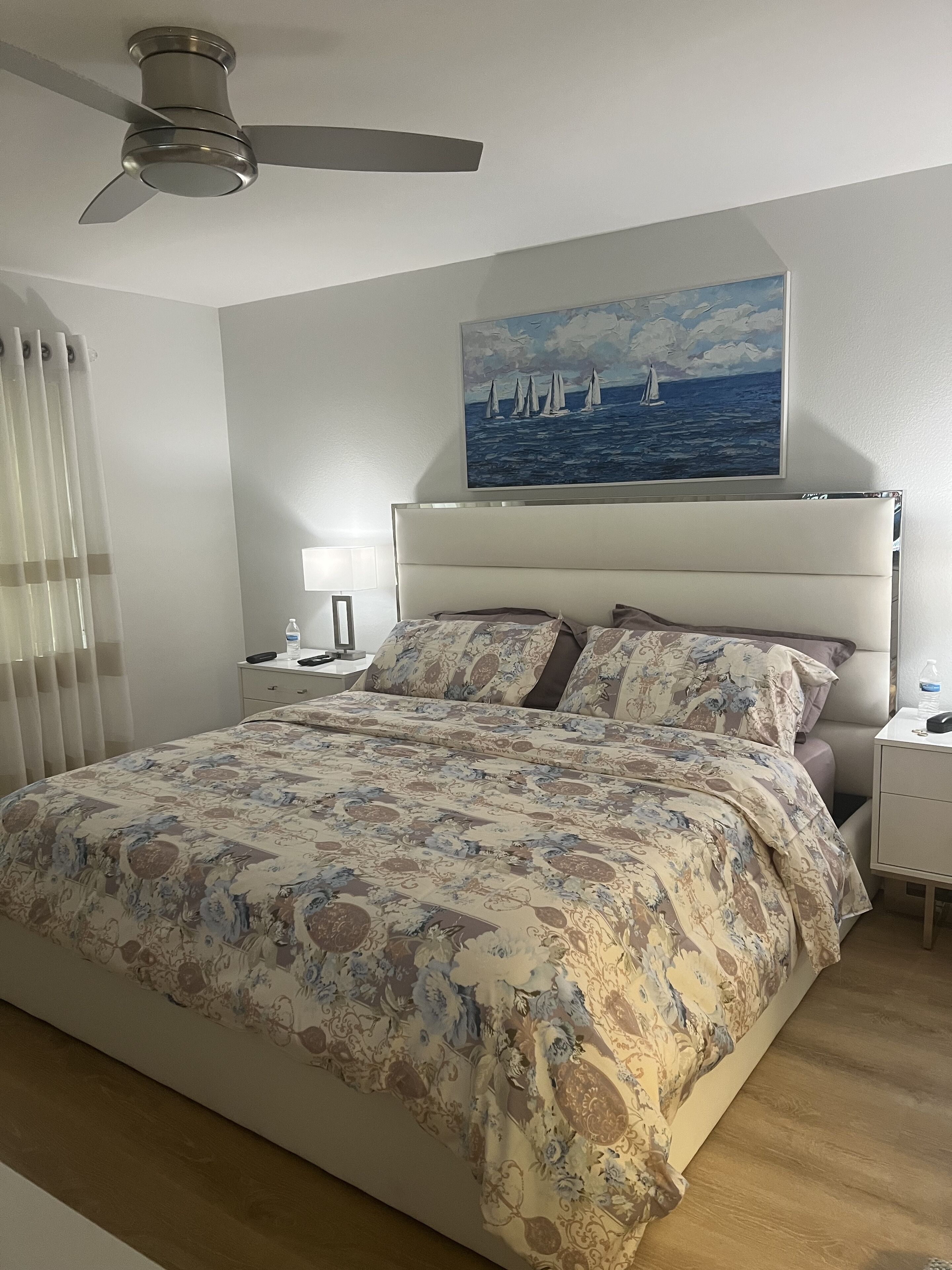 Et soveværelse med en seng, ventilator, natbord, lampe og et maleri af sejlbåde på væggen.