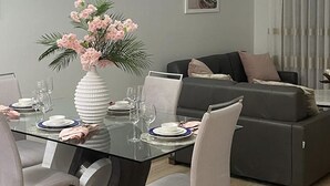 Un moderno comedor con una mesa de vidrio dispuesta para cuatro personas, rodeada de sillas grises tapizadas. Un jarrón blanco con flores rosas está sobre la mesa.