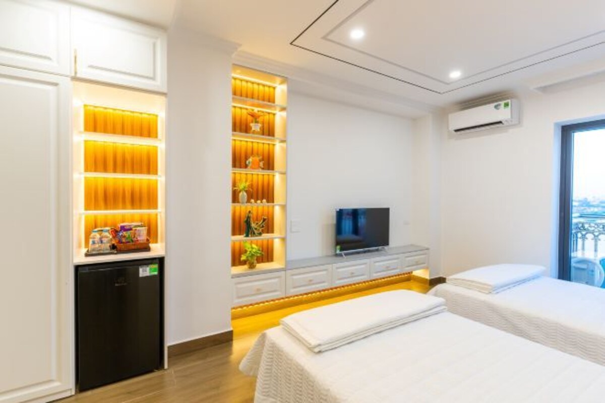 Une chambre d’hôtel équipée d’un lit, d’une télévision, d’un mini-réfrigérateur et d’une étagère intégrée, le tout éclairé d’une lumière chaleureuse.