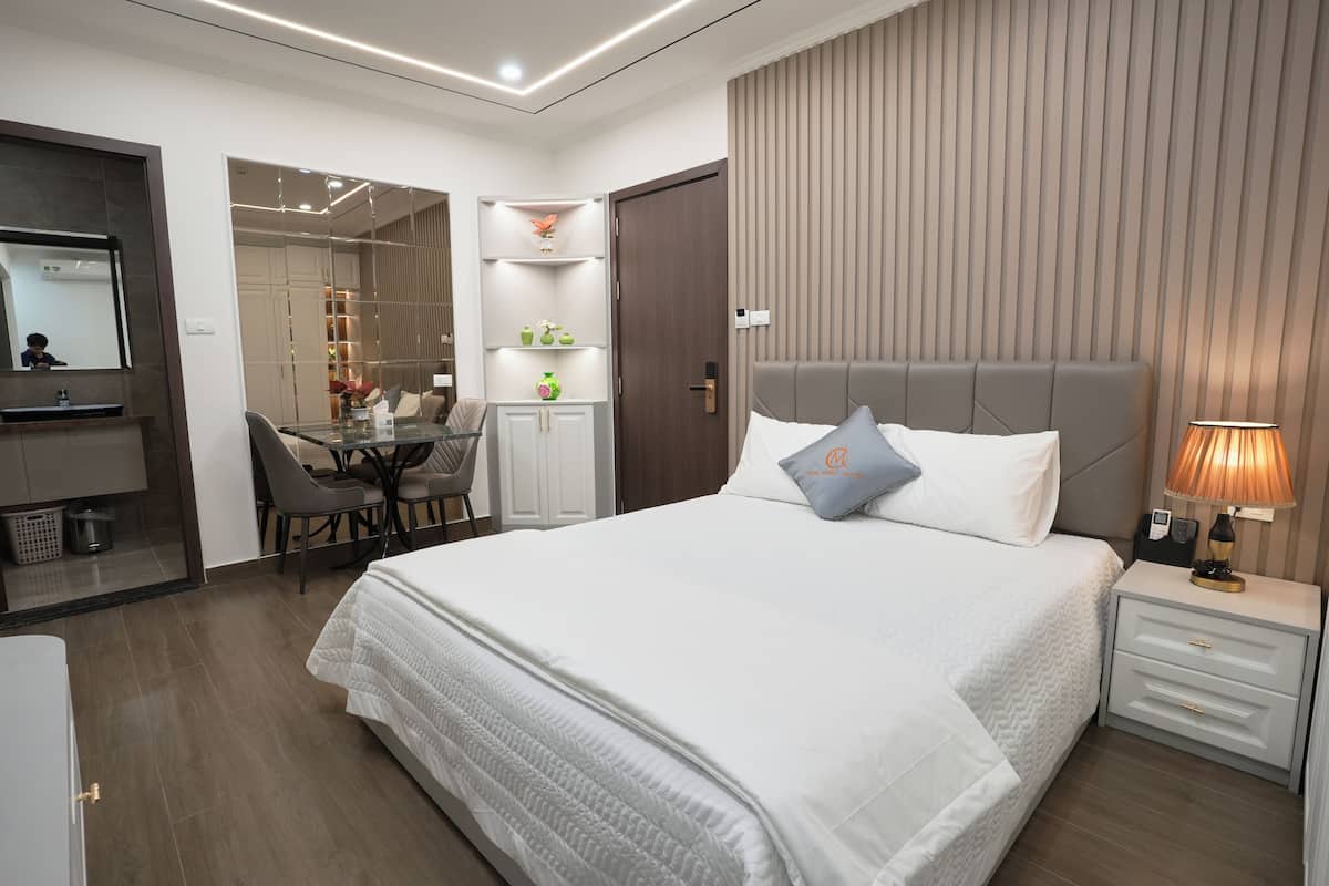 A modern hotel room with a large bed, bedside table, and a desk with chairs.