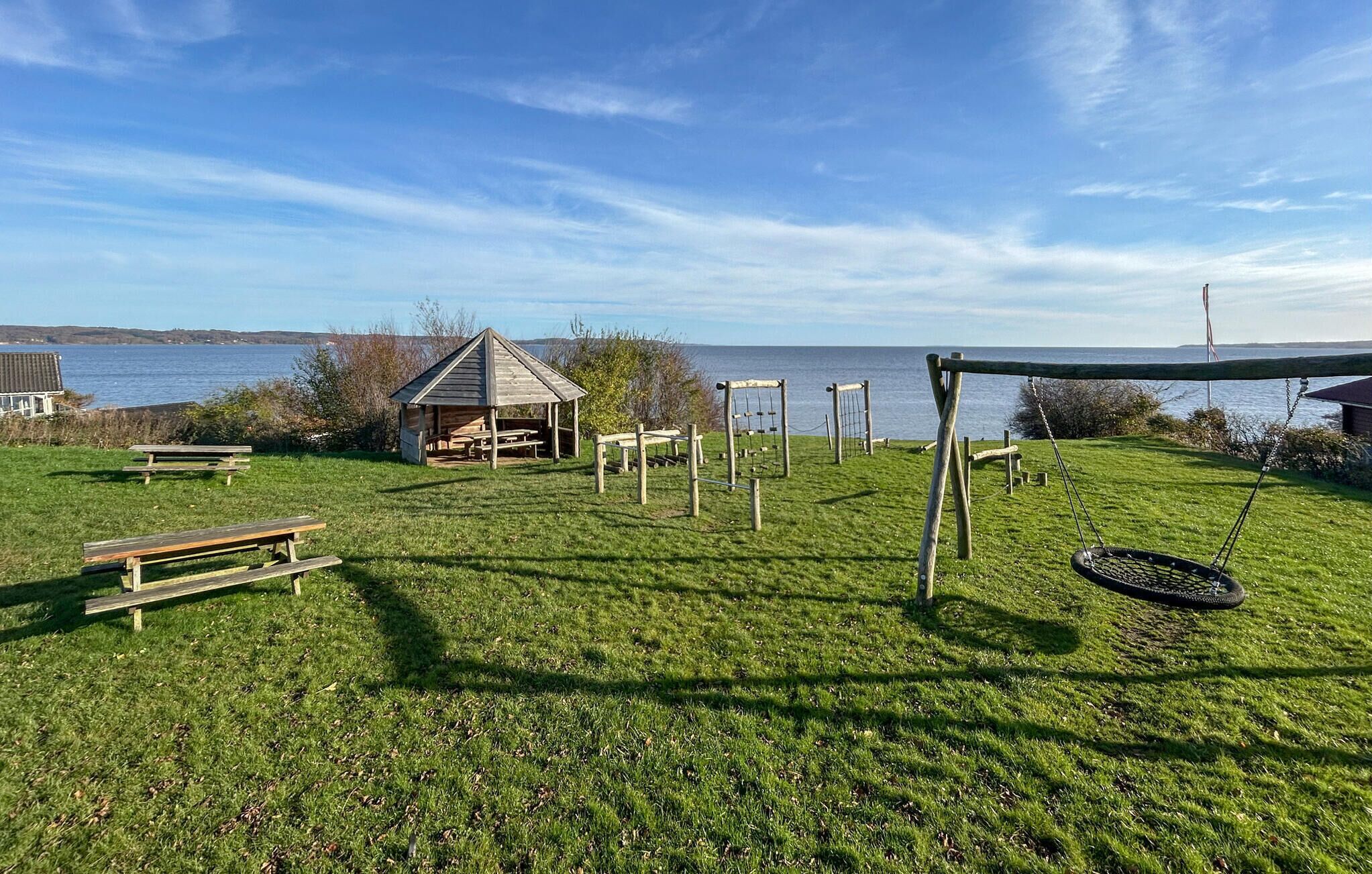 Un espacio al aire libre con césped, mesas de picnic, un conjunto de columpios y un pequeño refugio junto al agua.