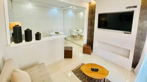 Een moderne woonkamer met een bank, een salontafel, een aan de muur bevestigde televisie en een eethoek, zichtbaar door een glazen scheidingswand.