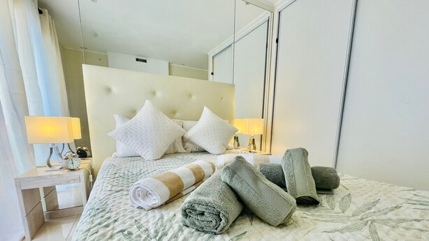 Een hotelkamer met een groot bed, twee bedlampjes, een nachtkastje met een klok en een spiegel aan de muur.