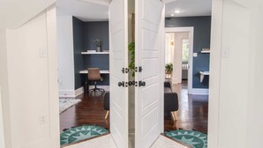 Un couloir avec une porte blanche donnant sur une pièce où se trouvent un bureau et une chaise.