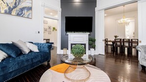 Un salon moderne avec un canapé bleu, un téléviseur à écran plat fixé au-dessus d’une cheminée, et un coin repas visible à travers une porte ouverte.