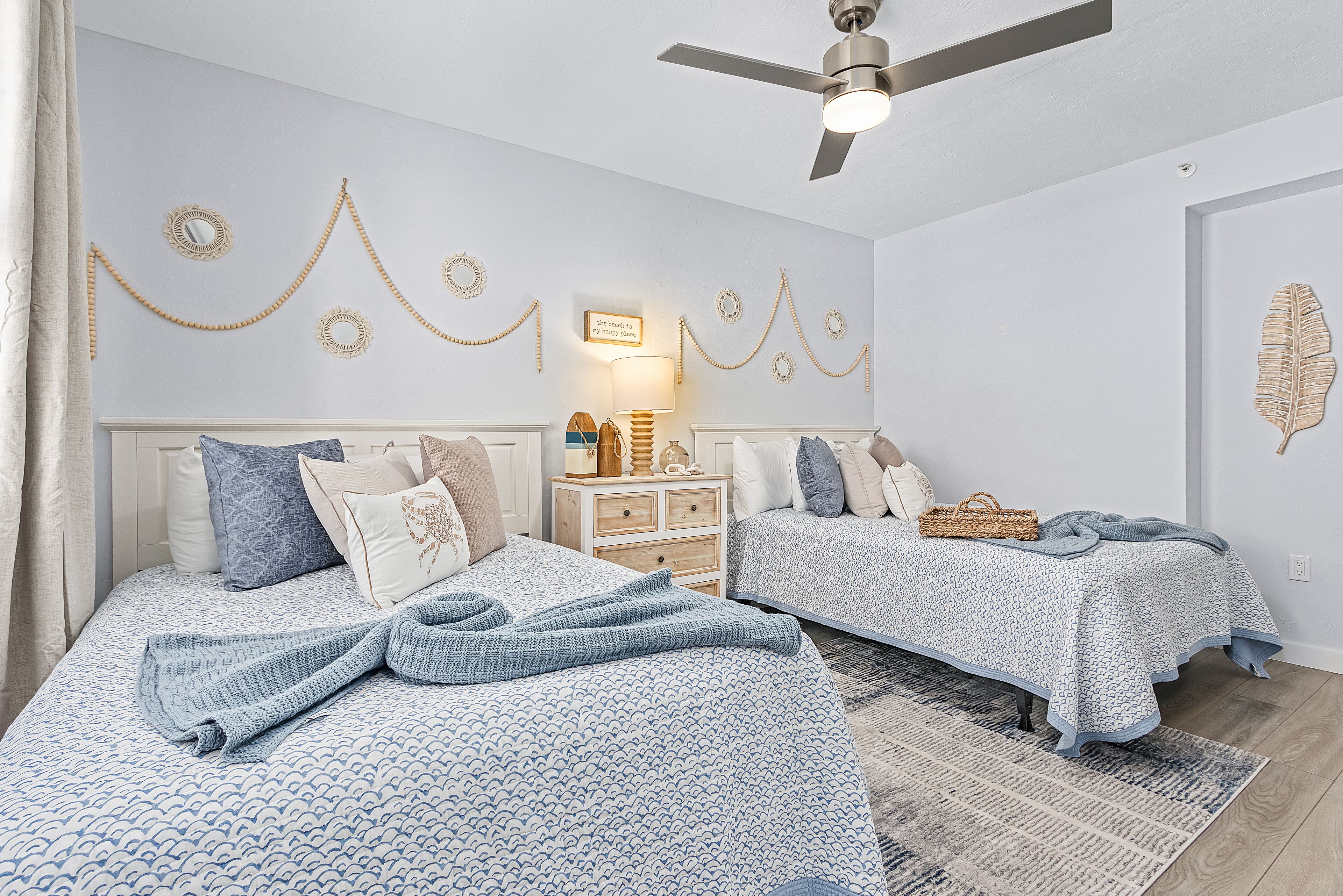 A bedroom with two beds, a ceiling fan, wall decorations, and a bedside table with a lamp.
