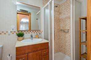 A bathroom with a white sink, wooden vanity, a mirror, a shower with a tiled wall, and a small plant on the counter.