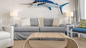 Un salon avec un canapé gris, deux tables d’appoint avec des lampes, une table basse en osier et une grande sculpture de poisson accrochée au mur.