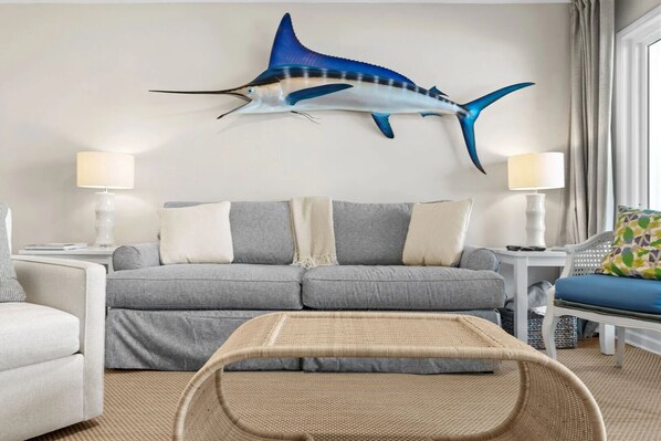 A living room with a grey sofa, two side tables with lamps, a wicker coffee table, and a large wall-mounted fish sculpture.
