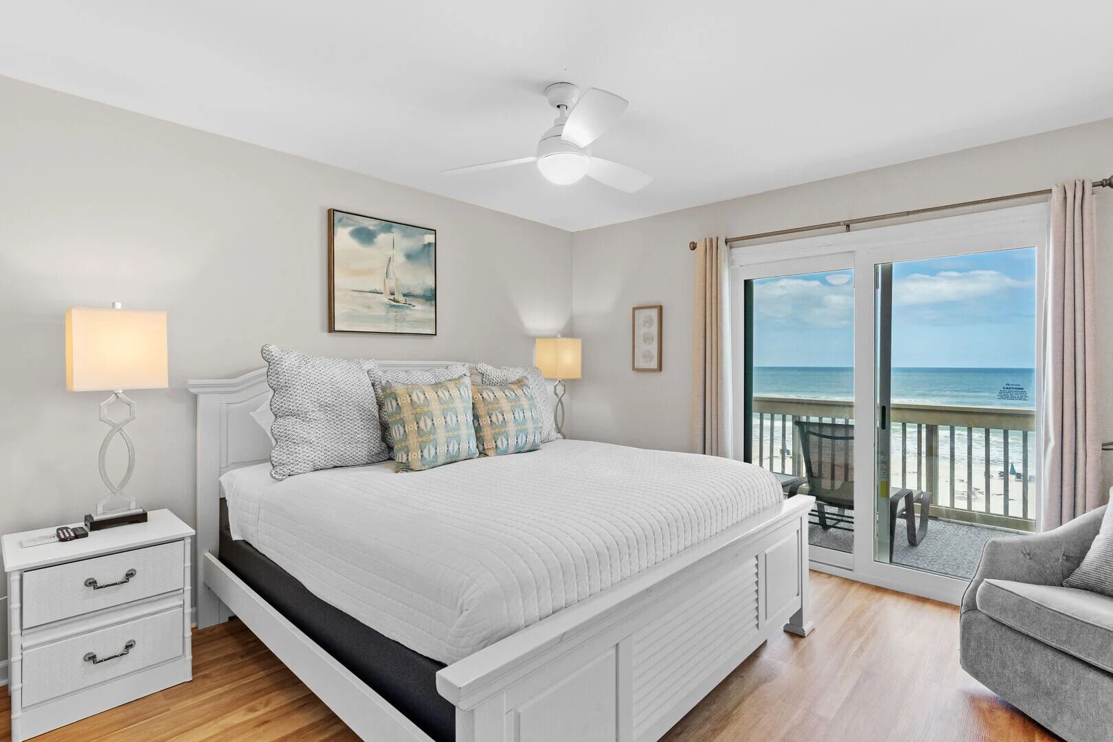 A bedroom with a bed, bedside tables, a ceiling fan, and a view of the beach through a sliding glass door.