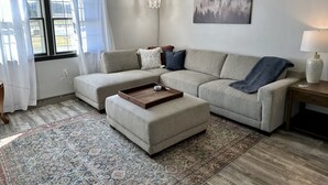 Una sala de estar con un sofá modular, una mesa de centro, un tapete, un cuadro en la pared y una lámpara sobre una mesita auxiliar.