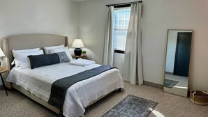 Un dormitorio ordenado con cama, lámparas de noche, una ventana con cortinas, un espejo y una alfombra.