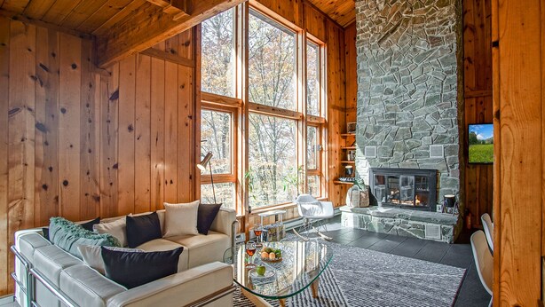 A cozy cabin interior with a stone fireplace, wooden walls, and a large window overlooking a forest. - 3 Br: N Westridge 222-7 Alpenglow: 3BR,2 Bath (Snowshoe)