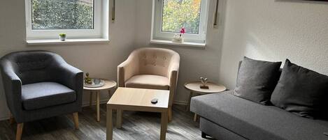 Un salón con un sofá gris, un sillón gris, una mesa de centro de madera y dos ventanas con persianas.
