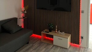 A modern living room with a flat-screen TV mounted on a wooden panel wall, a dark sofa, a small wooden coffee table with a plant, and red LED strip lighting along the ceiling. - Gartenparadies Apartment  in Urbar (Urbar)