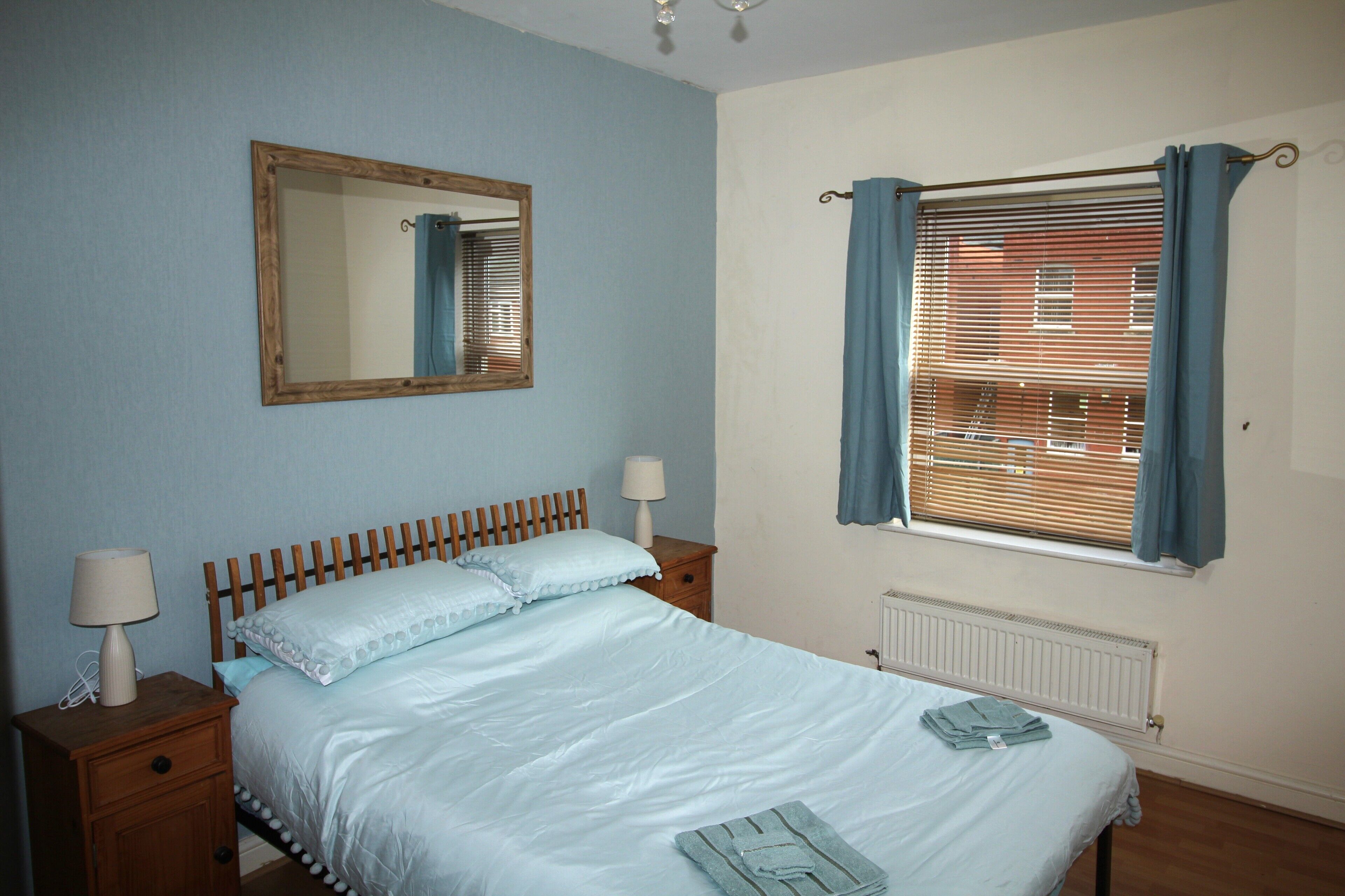 A bedroom with a wooden bed, two bedside tables with lamps, a mirror, a window with curtains, and a radiator.