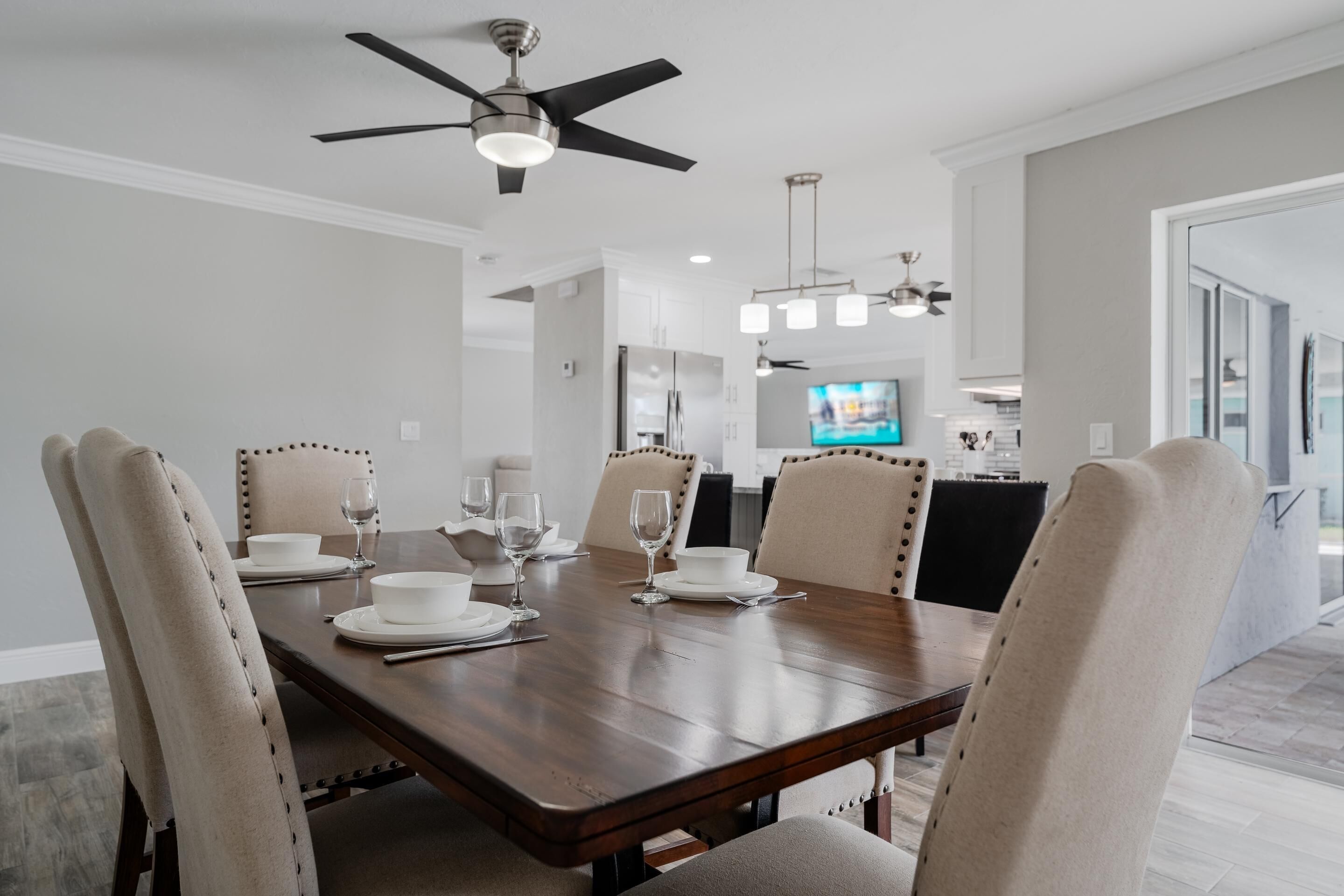 Un coin repas avec une table en bois dressée pour six personnes, des chaises à revêtement beige, un ventilateur de plafond, des lampes suspendues et un téléviseur en arrière-plan.