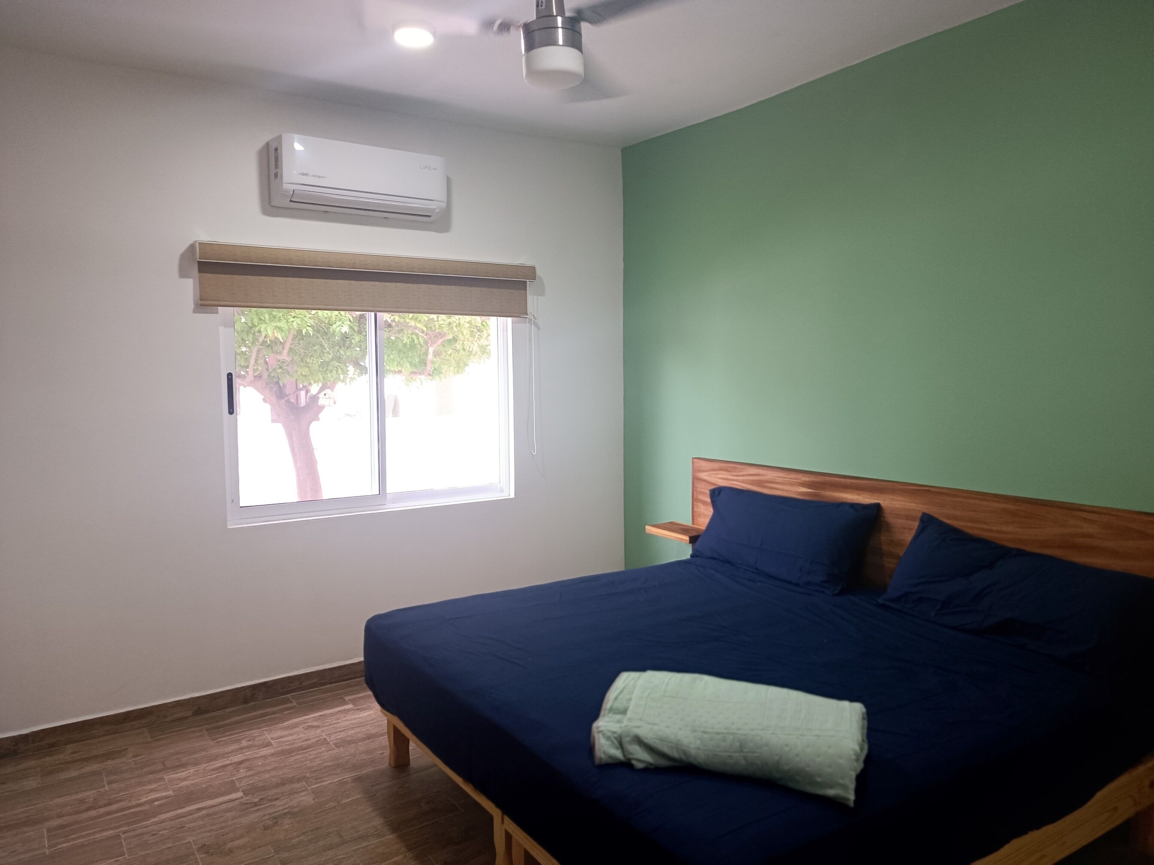 Une chambre à coucher avec un lit, une fenêtre donnant sur des arbres, un climatiseur et un ventilateur de plafond.