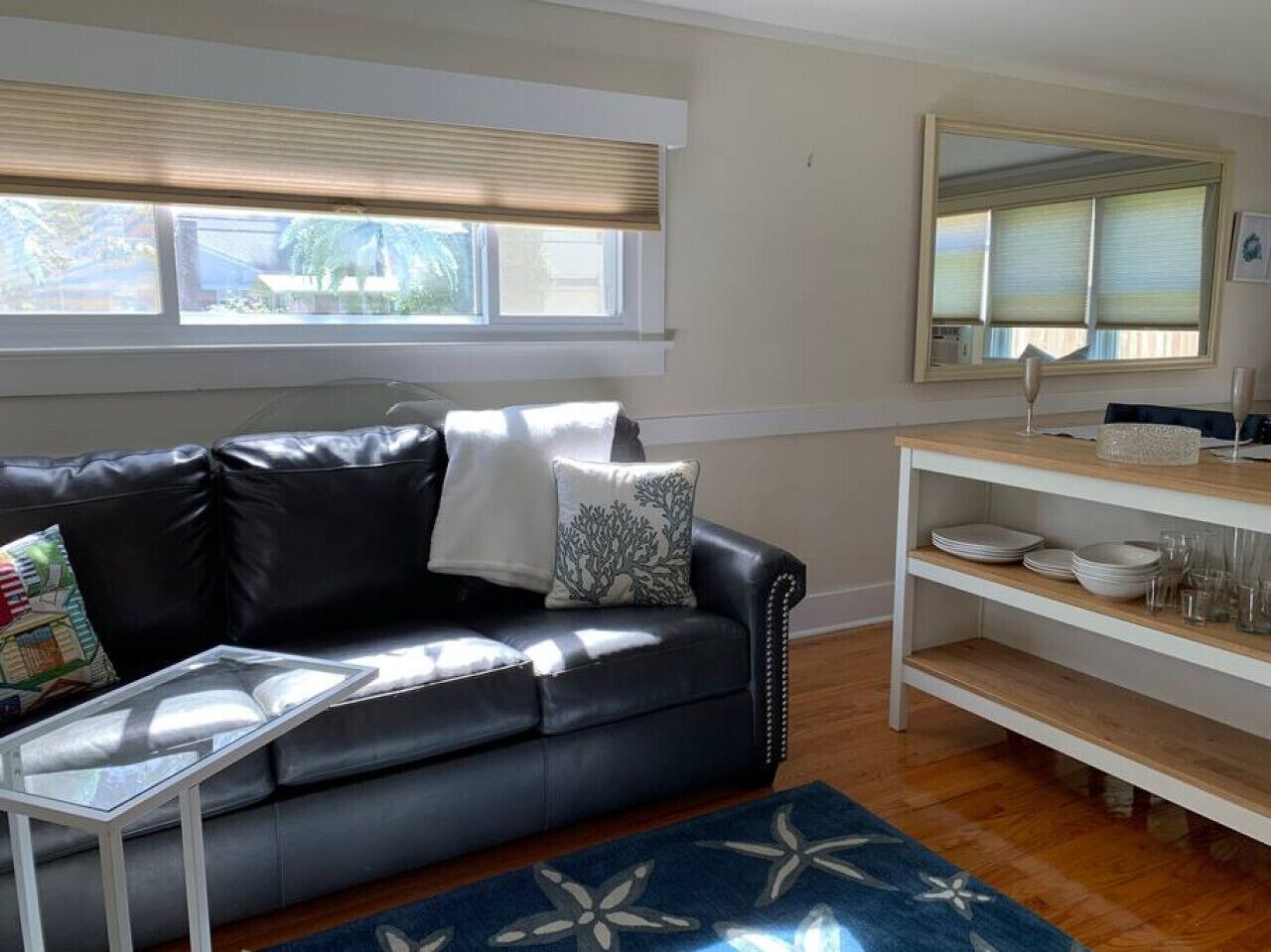 A living room with a black leather sofa, a white coffee table, a blue patterned rug, a white shelving unit with dishes, and a large mirror on the wall.