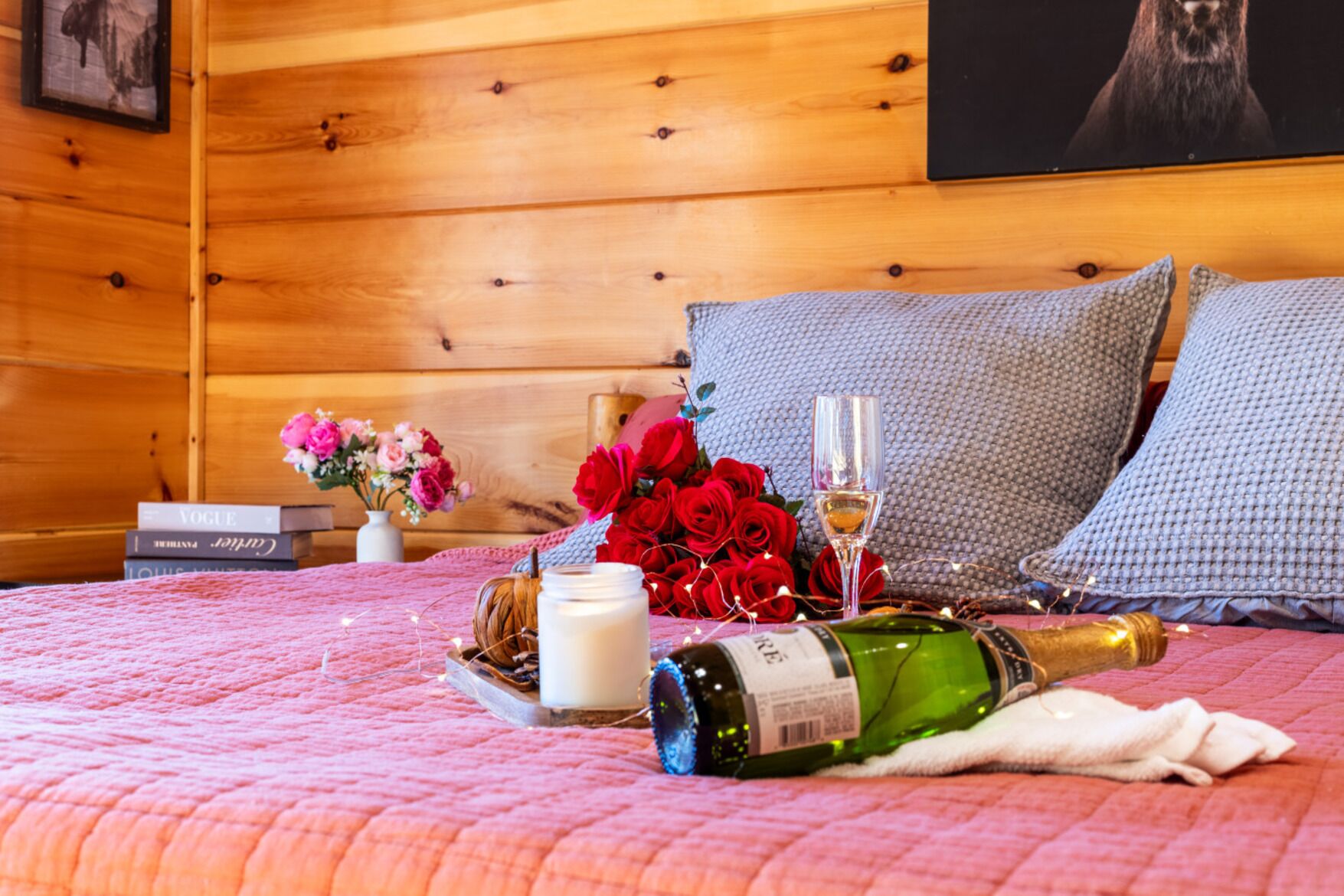 Una habitación acogedora con cabecera de madera, cama con colcha rosa, un ramillete de rosas rojas, una botella de champán y una copa de vino.