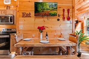 A wooden interior with a dining table set for four, a TV mounted on the wall, and a kitchen area with appliances.