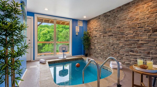 A pool area with a basketball hoop, a stone wall, and a view of greenery outside.