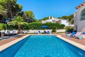 A rectangular swimming pool with clear blue water, surrounded by lounge chairs and umbrellas, with a white building and greenery in the background. - Villa La Tranquilidad - Plus (Benissa)