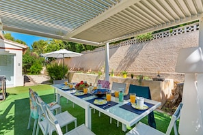 An outdoor dining area with a table set for two, surrounded by chairs, under a covered patio with a stone wall backdrop. - Villa La Tranquilidad - Plus (Benissa)