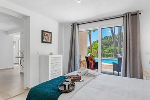 A bedroom with a bed, a bedside table, a window with a view of a pool and greenery, and a door leading to another room. - Villa La Tranquilidad - Plus (Benissa)