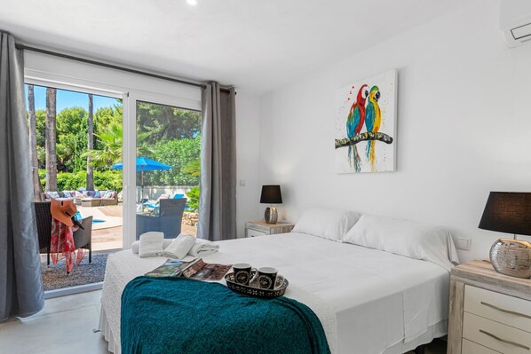 A hotel room with a bed, a nightstand, a lamp, a painting of parrots, and a view of an outdoor area with a pool and lounge chairs.