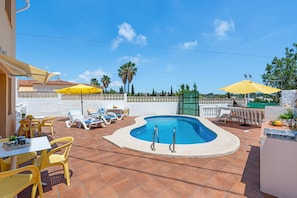 A swimming pool with sun loungers and umbrellas, surrounded by a white wall and palm trees.