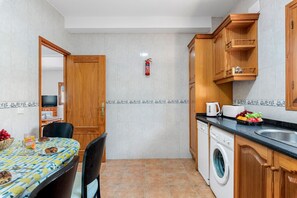 A compact kitchen with a washing machine, a dining table with chairs, and a small television in the background.