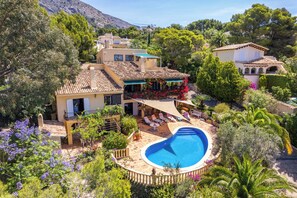 Una villa in stile mediterraneo con piscina, circondata da vegetazione rigogliosa e uno sfondo montuoso.