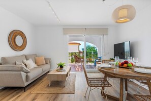 Una sala de estar moderna con sofá, mesa de centro, mesa de comedor y sillas. Hay un televisor y una zona de comedor con fruta y vasos.