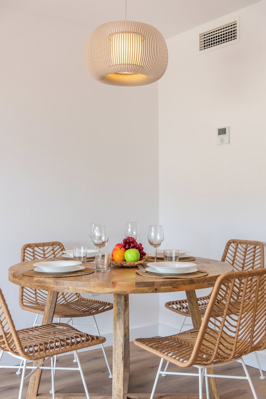 Una mesa de madera redonda con cuatro sillas de mimbre, una lámpara colgante tejida y un frutero sobre la mesa.