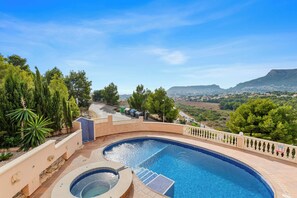 A swimming pool with a curved design, surrounded by a low wall and a railing, with a view of a hilly landscape in the background.