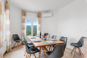 A dining area with a wooden table set for six, surrounded by black chairs with wooden legs, a window with curtains, and an air conditioning unit on the wall.