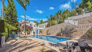 A swimming pool with a stone wall and lounge chairs, surrounded by greenery and a clear sky.