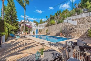 A swimming pool with a stone wall and lounge chairs, surrounded by greenery and a clear sky.