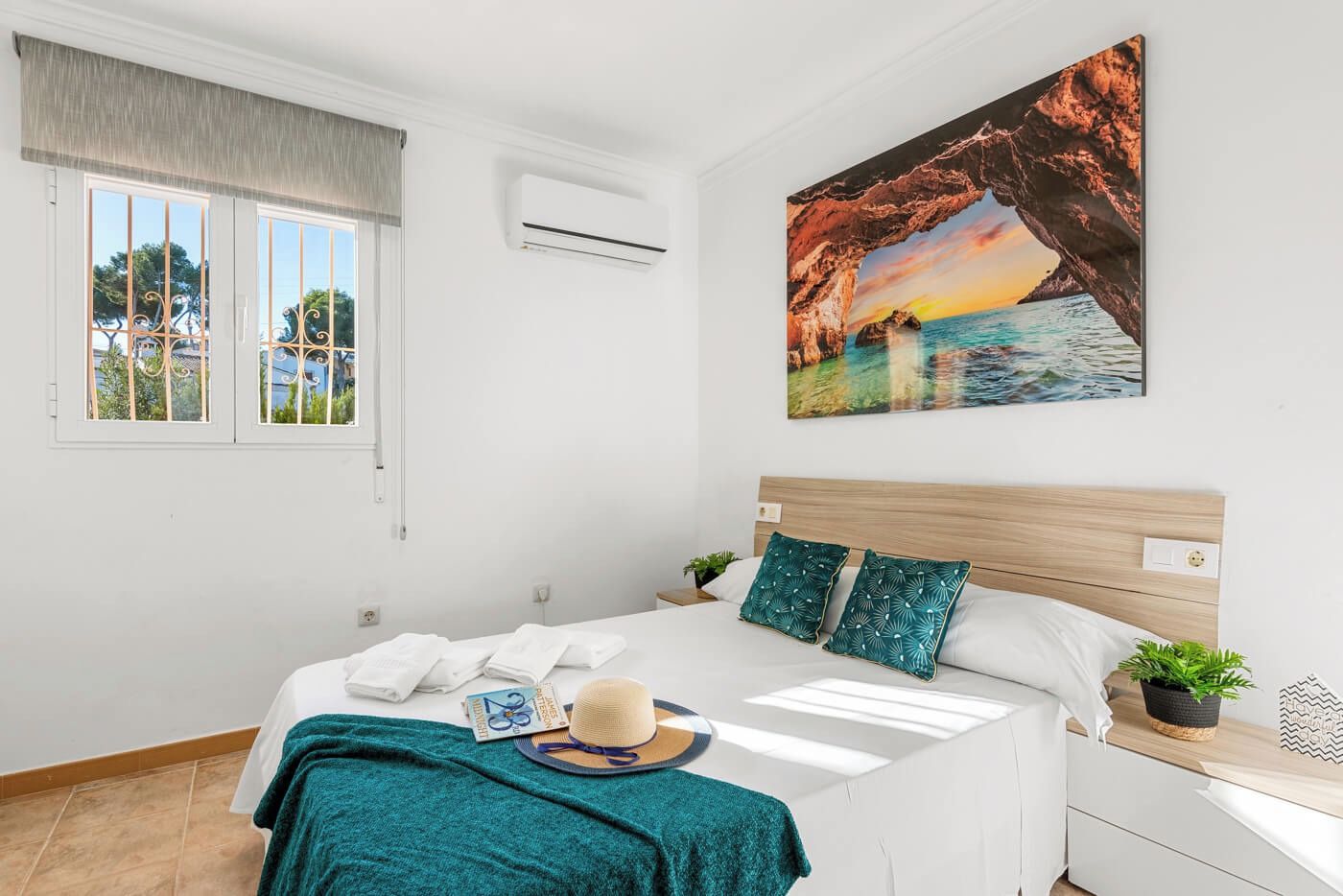 A bedroom with a bed, a window, an air conditioning unit, a wall-mounted painting, and a bedside table with a plant.
