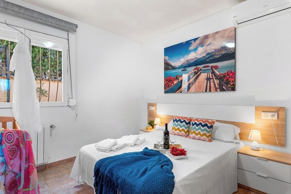 A bedroom with a bed, a window with curtains, a painting of a scenic landscape, and a bedside table with a lamp.