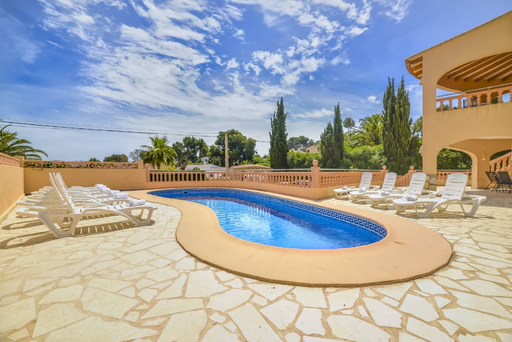 A swimming pool with lounge chairs, surrounded by a stone patio and a building with a balcony.