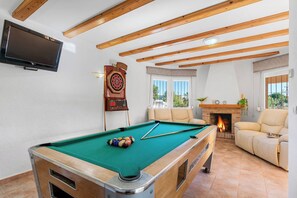 A pool table with balls, a dartboard, a television, a fireplace, and a sofa in a room with wooden beams.